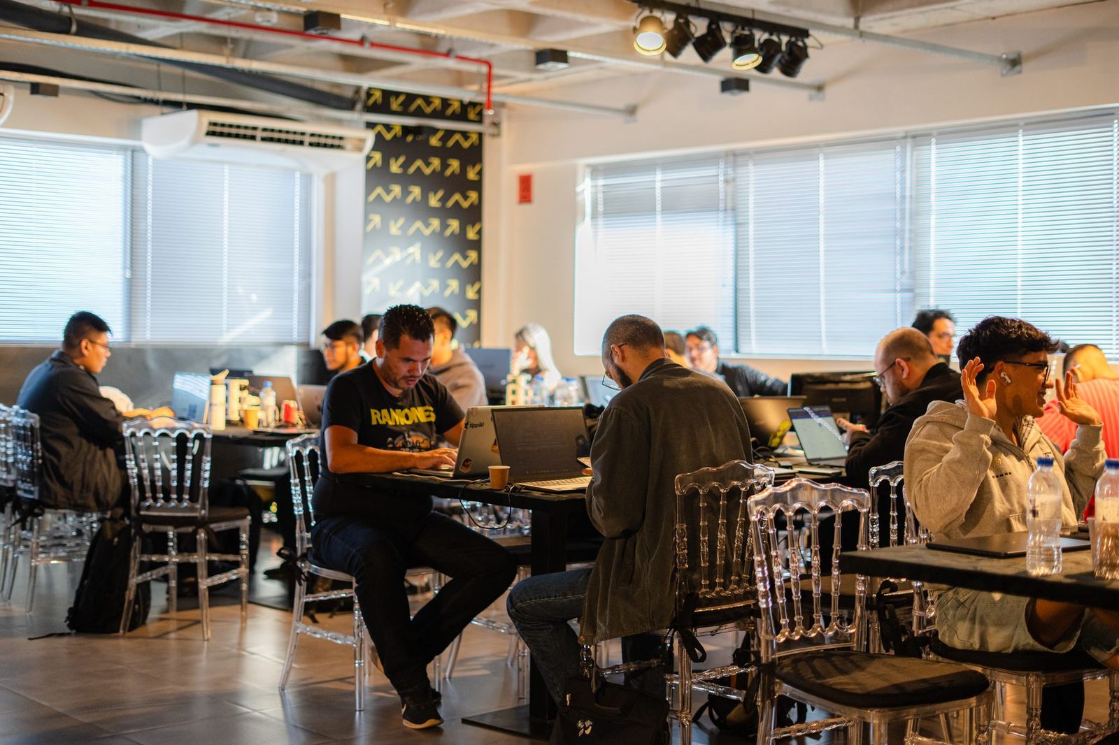 Coworking room lit by natural light with participants working on laptops at tables, transparent chairs and a black panel with yellow arrows in the background.