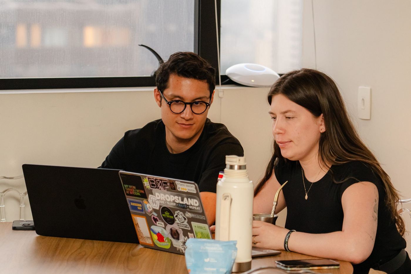 Two builders side by side looking at the same sticker-covered laptop, on a wooden table with a thermos and a mate gourd.