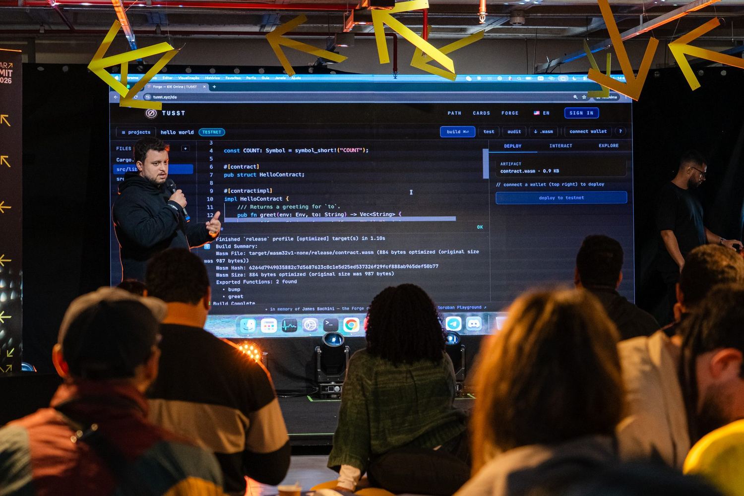 Main stage with a presenter at the microphone in front of a giant LED panel showing an IDE with Rust/Soroban code and the build log of a contract being deployed to testnet.