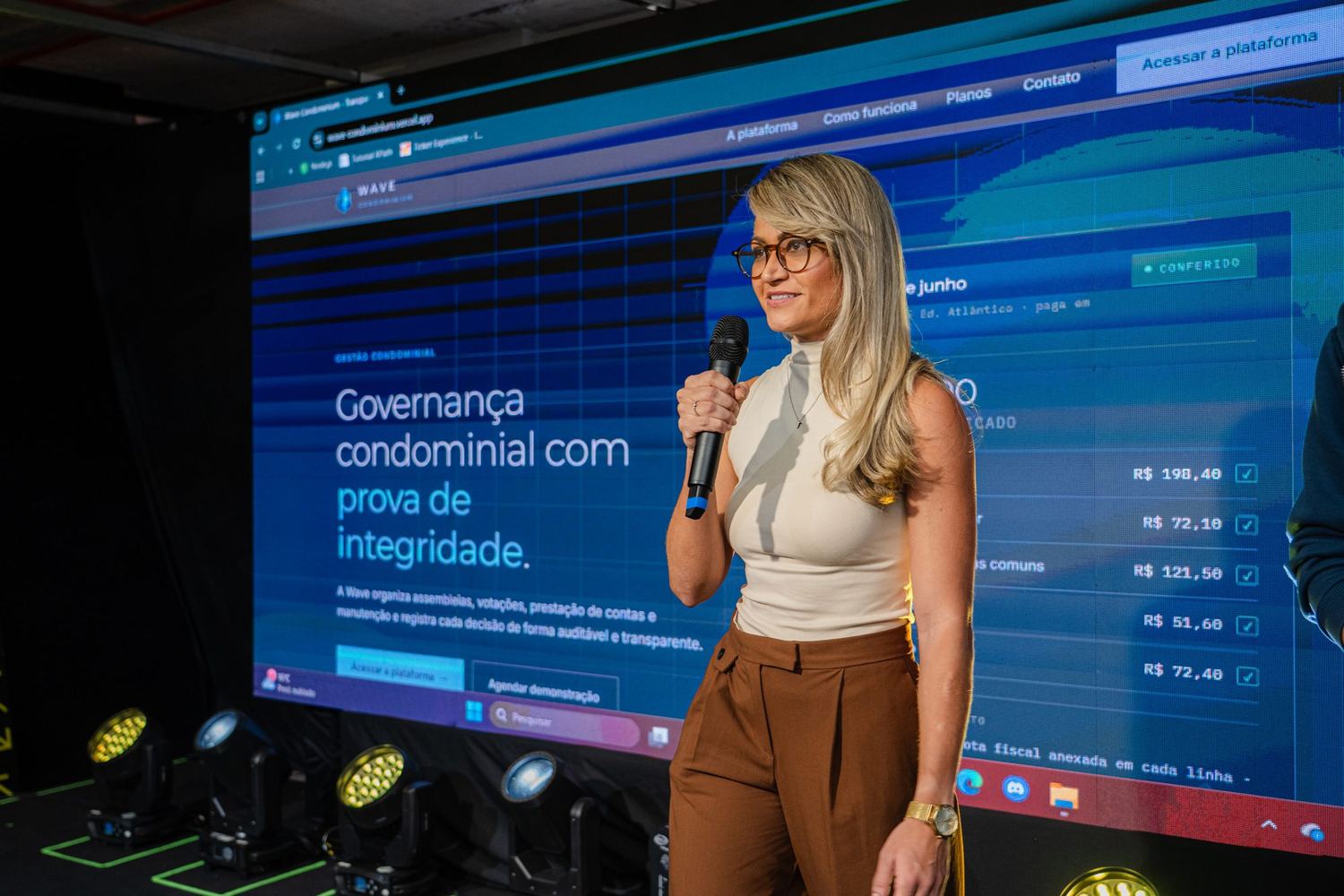 Presenter on the showcase stage with a microphone; behind her, an LED screen displays the page of a condominium-governance product with values in reais.